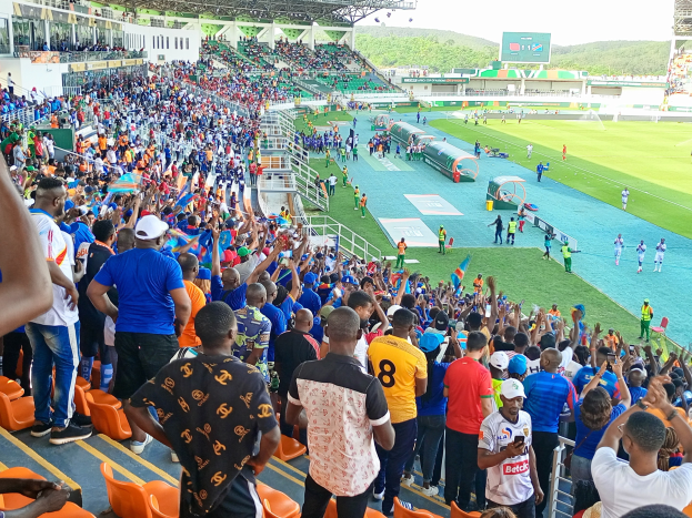Eine große Menschenmenge versammelt sich vor einem eingezäunten Fußballfeld, wobei einige auf Stühlen sitzen und andere stehen, während Hügel und ein klarer blauer Himmel den Hintergrund bilden.