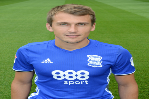 Ein Mann in einem blauen T-Shirt sitzt auf einem Fußballfeld und posiert mit einem selbstbewussten Ausdruck, wahrscheinlich ein Flügelspieler.