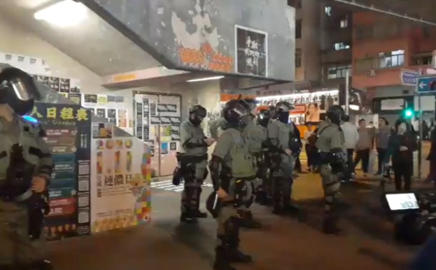Polizeibeamte in Einsatzkleidung stehen vor einer Menge von Helikopterträgern mit Waffen, mit Plakaten an Wänden, Deckenleuchten, Gebäuden und einer Ampel im Hong Kong.