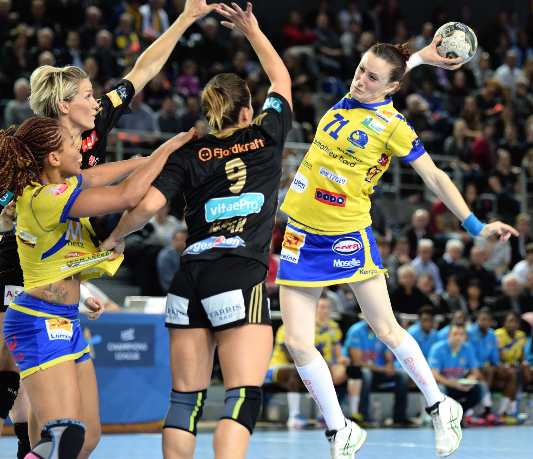 Eine Gruppe von Frauen beim Handballspielen, wobei eine den Ball hält, während unscharfe Zuschauer im Hintergrund sitzen.