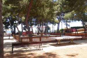 Ein Park mit einem zentralen Tischtennis-Tisch, umgeben von Bänken, einem Zaun, Schaukeln, Pfosten, Pflanzen und Bäumen, vor einer Wasserfläche und einem klaren blauen Himmel.