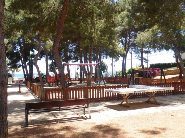 Ein Park mit einem zentralen Tischtennis-Tisch, umgeben von Bänken, einem Zaun, Schaukeln, Pfosten, Pflanzen und Bäumen, vor einer Wasserfläche und einem klaren blauen Himmel.