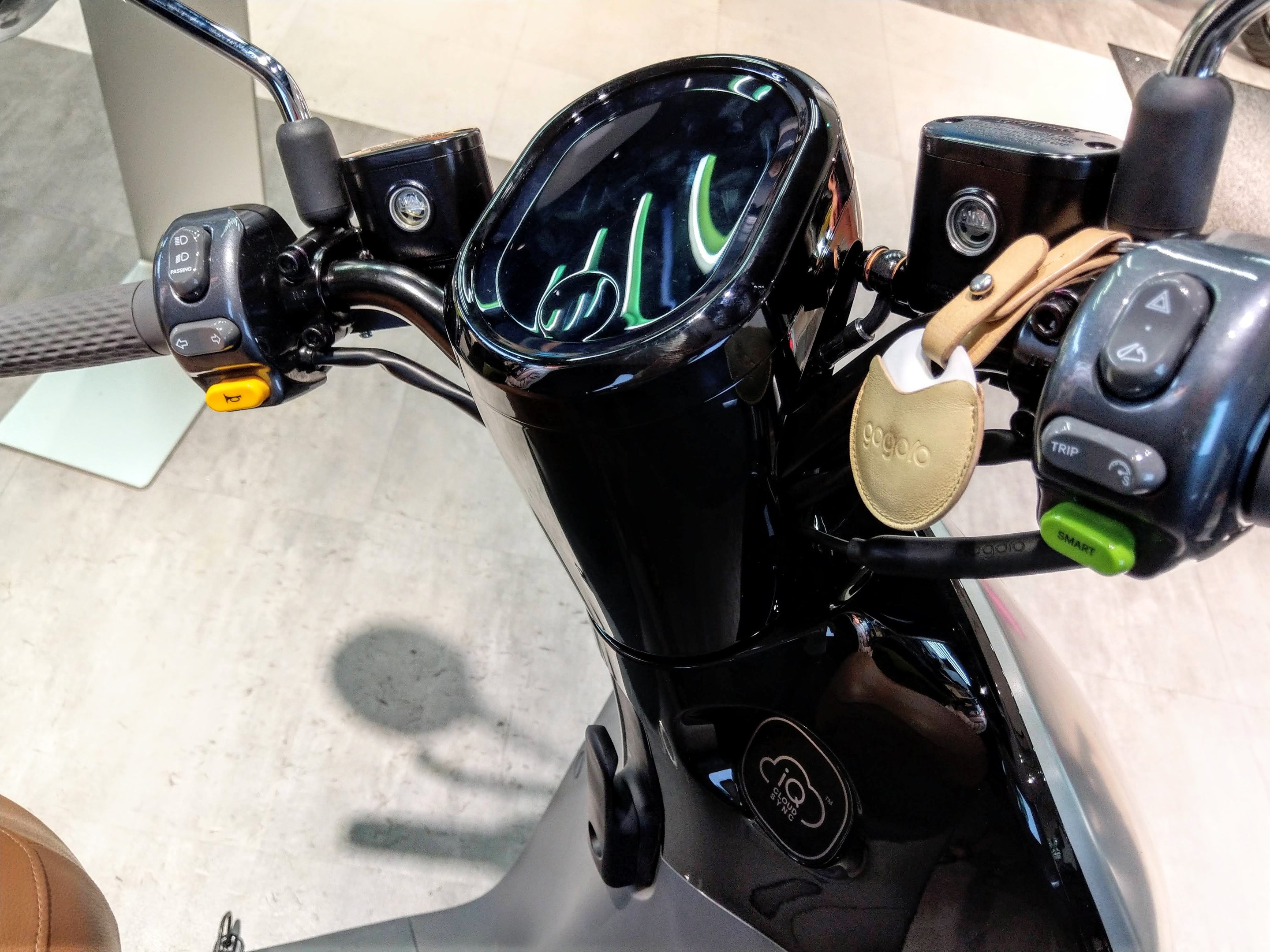 Nahaufnahme eines schwarzen E-Scooters auf einer Ausstellung, mit Lenker und Sitz, sowie einer Säule und einem Boden im Hintergrund.