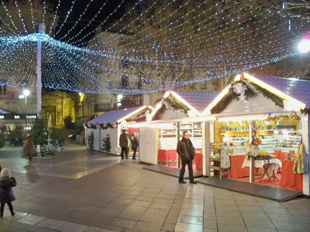 Ein nächtlicher Weihnachtsmarkt in einer Stadt, mit Menschen, die stehen, sitzen und ein Kind halten, umgeben von festlichen Lichtern, Pflanzen, Bäumen und Gebäuden mit Fenstern, unter einem bewölkten Himmel.