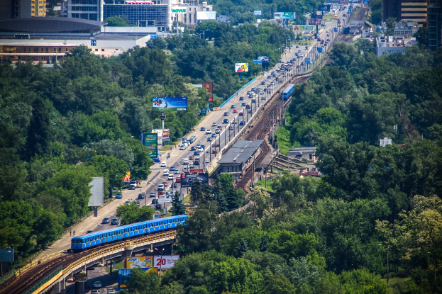 Eine belebte Straße mit Verkehr, ein Zug auf Höhenbahn, umgeben von Bäumen, Gebäuden, Plakaten, Mästen und Eisenbahninfrastruktur.