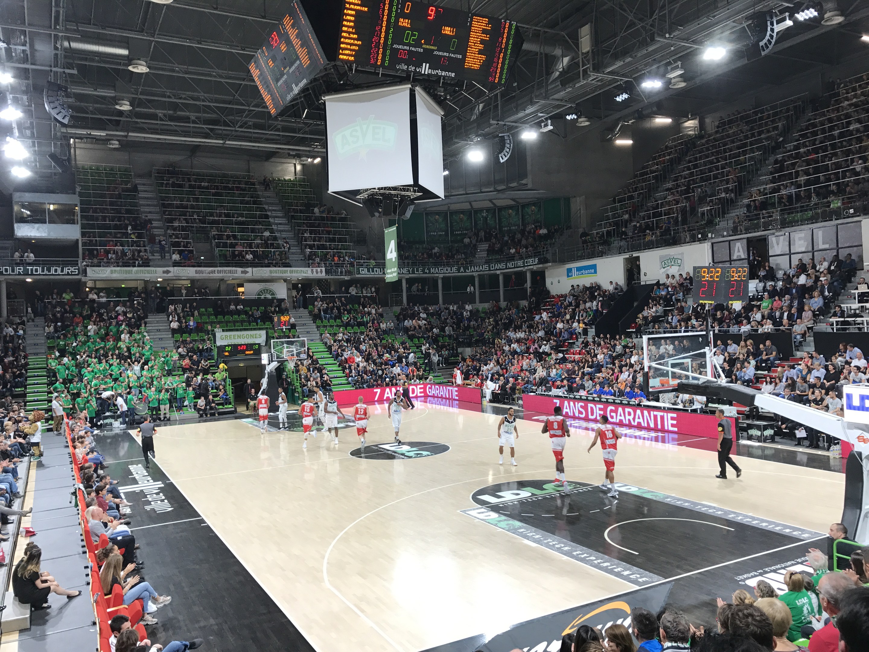 Basketballspiel in einer großen Arena mit Publikum, Anzeigetafeln mit "Asvel vs Galatasaray" und Deckenbeleuchtung und -bildschirmen.