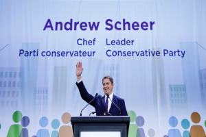 Andrew Scheer, der Vorsitzende der konservativen Partei, steht vor einem Podium mit zwei Mikrofonen und einem Bildschirm, mit einem Banner im Hintergrund, das Text und Gebäudeabbildungen zeigt.