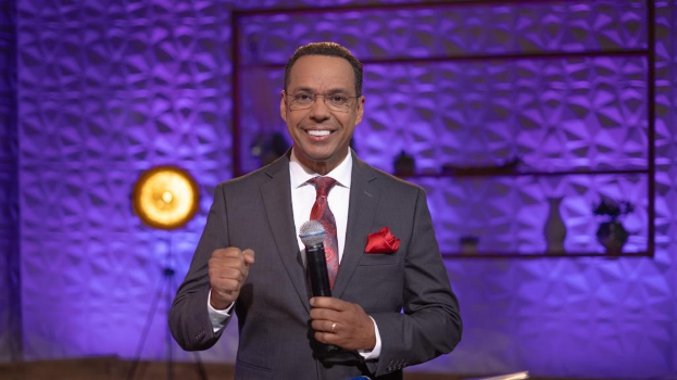 Stephen Colbert, Moderator von 'The Daily Show with Trevor Noah', steht auf einer Bühne und hält ein Mikrofon in der Hand, trägt einen Anzug, Krawatte und eine Brille, mit einem Scheinwerfer und Ständer hinter ihm.
