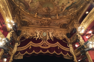 Theaterinnenraum mit roten Vorhängen, Kronleuchtern, Deckenmalerei und einer Bühne mit ein paar Menschen davor.