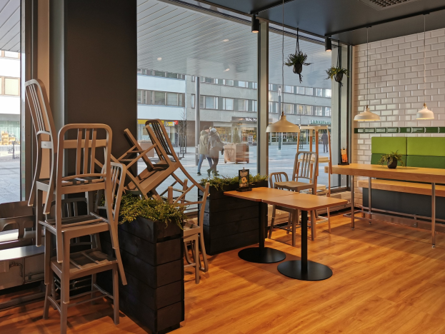 Ein Restaurant in Berlin mit Holztischen und -stühlen, umgeben von Pflanzen und Lichtern, mit einem großen Fensterblick auf Menschen, die auf der Straße gehen, Gebäude und den Himmel.