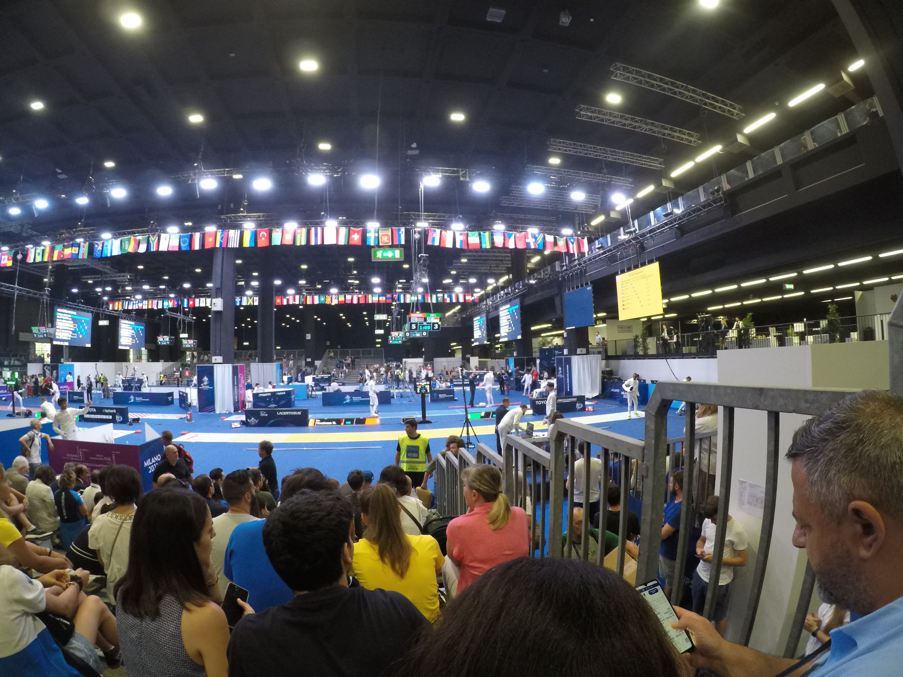 Eine große Menschenmenge sitzt und steht in einer Indoor-Sportarena mit Bildschirmen, Flaggen und Deckenleuchten im Hintergrund.