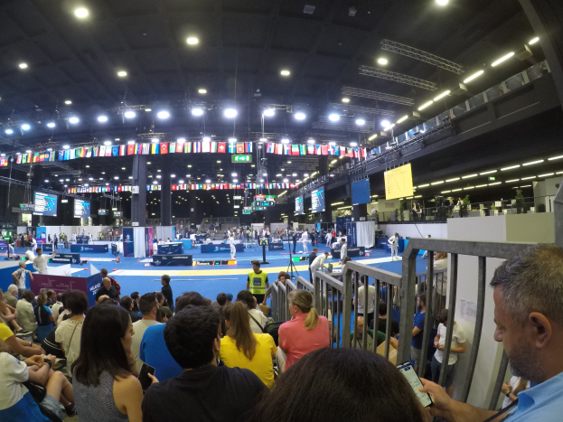 Eine große Menschenmenge sitzt und steht in einer Indoor-Sportarena mit Bildschirmen, Flaggen und Deckenleuchten im Hintergrund.