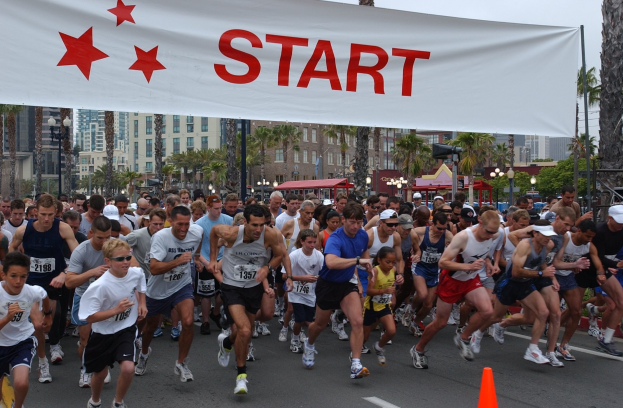Eine Gruppe von Menschen beim Laufen eines Marathons mit einem Verkehrskegel im Vordergrund und einer Fahne im Hintergrund, vor Bäumen, Laternenmasten, Gebäuden und einem klaren blauen Himmel.
