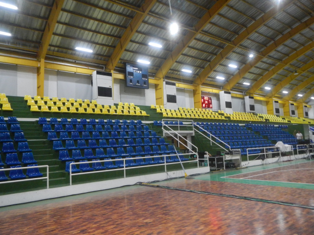 Großer Hallensportplatz mit Holzboden, blauen und gelben Sitzplätzen, Geländern, Treppen zum Sitzbereich, Wandbildschirmen, Deckenleuchten und einigen Personen.