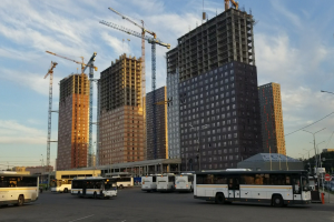Eine Gruppe von Bussen, die auf einem Parkplatz neben höhen Gebäuden geparkt sind, mit Straßeninfrastruktur und Bäumen im Hintergrund.