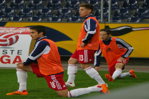 Drei junge Männer in Sportkleidung beim Fussballspielen auf einem Feld, mit Brettern, Stühlen und Pfählen im Hintergrund.