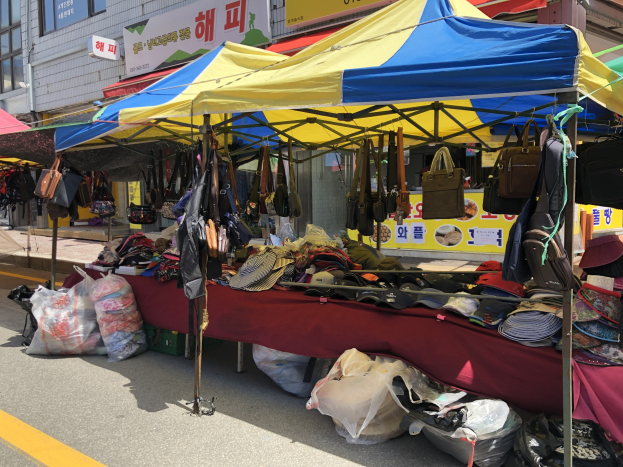 Ein Straßenmarkt mit Zelten, die Taschen, Hüte und andere Gegenstände präsentieren, entlang eines Bürgersteigs mit Gebäuden und Texttafeln im Hintergrund.