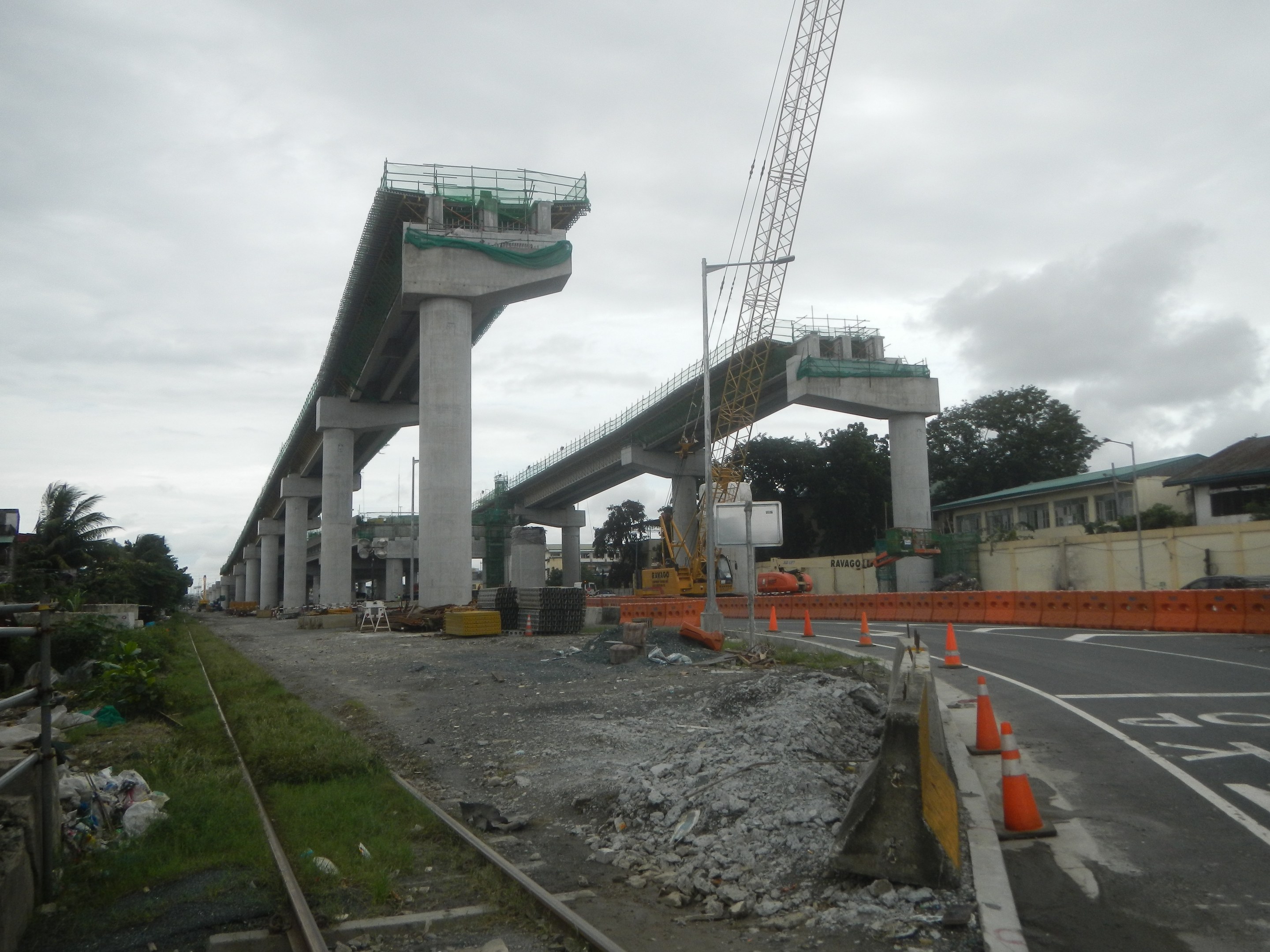 Baustelle mit einer Brücke im Hintergrund, Straße mit Verkehrskegeln markiert, Eisenbahnschiene links, verstreute Steine und Gras, Bäume und Gebäude säumen die Straße und ein bewölkter Himmel.