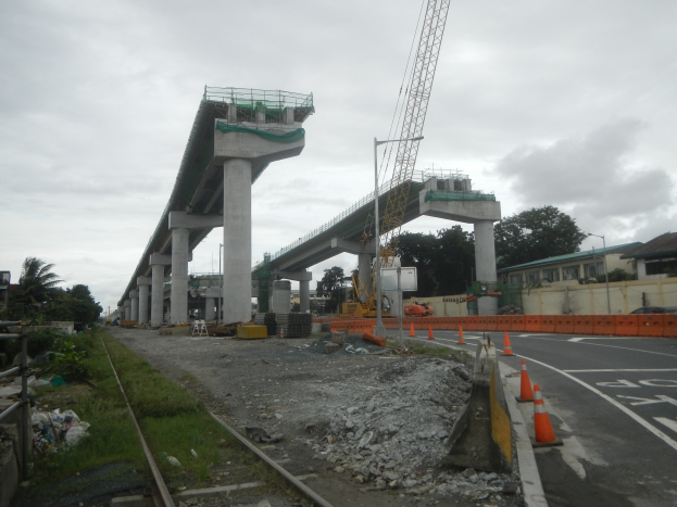 Baustelle mit einer Brücke im Hintergrund, Straße mit Verkehrskegeln markiert, Eisenbahnschiene links, verstreute Steine und Gras, Bäume und Gebäude säumen die Straße und ein bewölkter Himmel.
