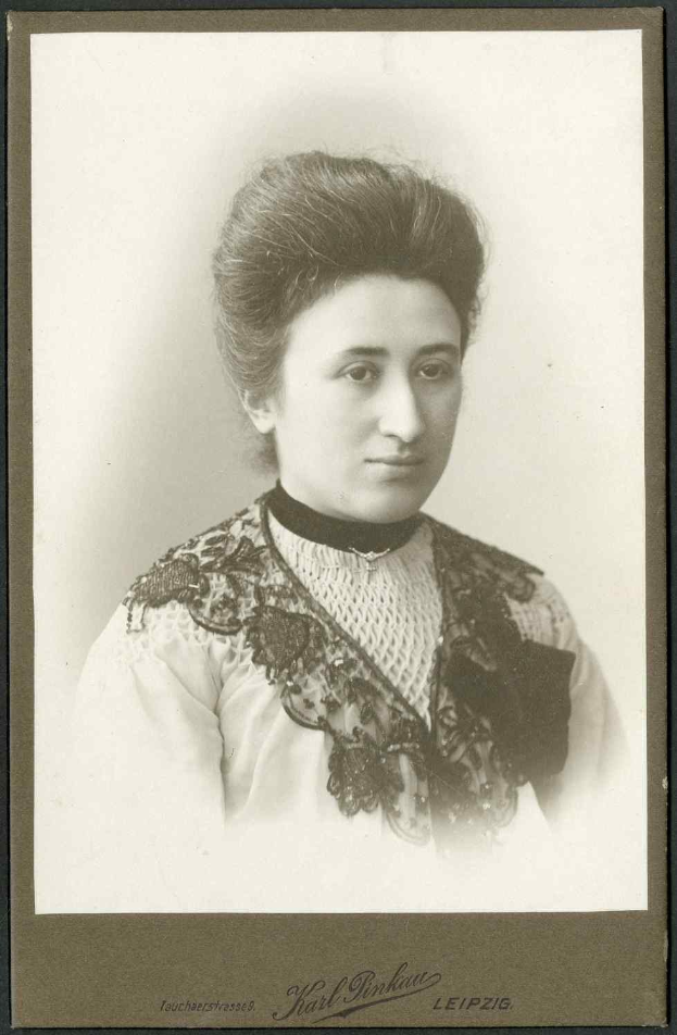 Ein Schwarz-Weiß-Foto einer Frau in einem weißen Kleid mit einem gemusterten Schultertuch, identifiziert als Karl Pinkau Leipzig, die direkt in die Kamera schaut.