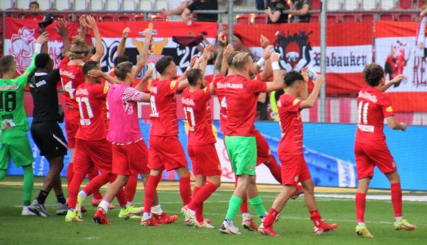 Gruppe von Fußballspielern, die ein Tor feiern, einige halten Flaschen oder klatschen, mit einem Zaun, Bannern, Metallstangen, Stühlen und einer Decke mit Deckenleuchten im Hintergrund.