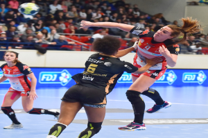 Eine Gruppe von Frauen spielt Handball auf einem Platz, während Zuschauer von Stühlen, Geländern und einer Wand im Hintergrund zusehen.