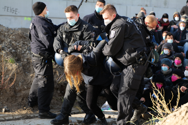 Gruppe maskierter Menschen in Mützen, einige mit Waffen, umringt eine Frau, die von der Polizei eskortiert wird, mit Pflanzen, Steinen und einer Wand mit Protesttext im Hintergrund.