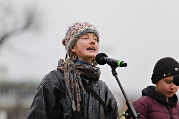 Eine Frau in einer schwarzen Jacke, einem Schal und einer Mütze singt in ein Mikrofon auf der Bühne, neben ihr eine Person in einer marinefarbenen Jacke und einer schwarzen Mütze, während eine unscharfe Menge zusieht.