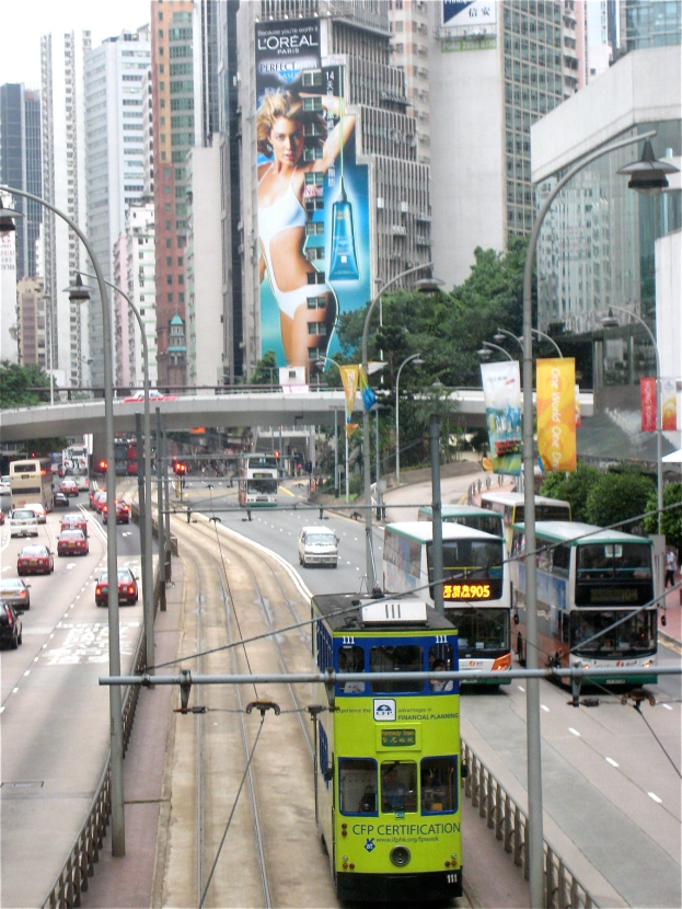 Lebendige Stadtstraße mit Doppeldeckerbussen, Autos und anderen Fahrzeugen, umgeben von hohen Gebäuden, Bäumen, Pfählen, Laternen, Schildern, Bannern und einer Brücke unter einem klaren blauen Himmel, mit Text unten.