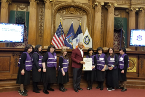 Eine Gruppe von Menschen steht vor Fahnen, ein Mann in der Mitte hält ein Zertifikat, flankiert von zwei Bildschirmen, vor einer hölzernen Wand mit kunstvollen Schnitzereien und Vorhängen bei einer Stadtratssitzung.
