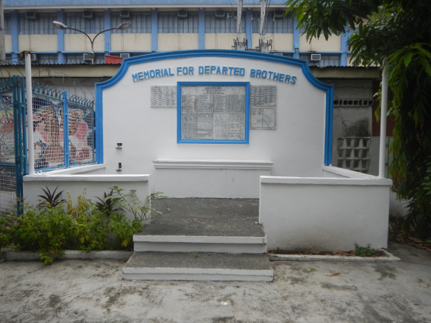 Gedenkstätte mit einer Gruppe von Pflanzen vor einem Gebäude mit einem blauen und weißen Schild, das einen Metallzaun, eine Treppe, Pfosten, Drähte, einen Baum und einen bewölkten Himmel zeigt.