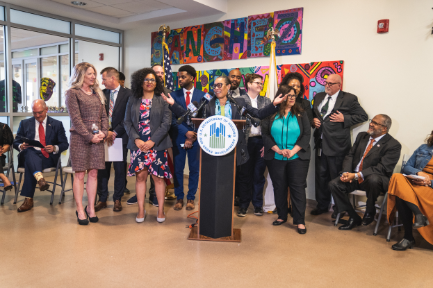 Eine Gruppe von Menschen, die in einem Raum mit einem Podium, einem Mikrofon und einer Tafel mit der Aufschrift "Sancho City Councilman Joins City Council" stehen und sitzen, mit Flaggen, einer Tafel, einem Feuerlöscher und Deckenleuchten an der Rückwand.