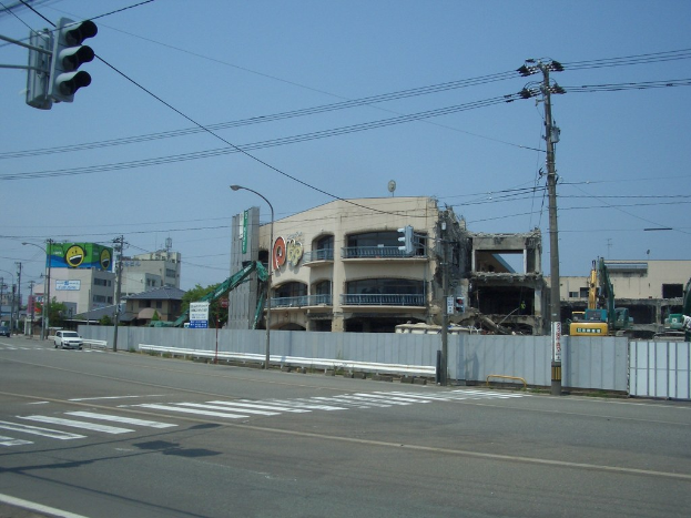 Eine Stadtstraße mit Fahrzeugen, Strommasten, Laternen und einer Ampel auf der linken Seite, mit Gebäuden, Bäumen und Himmel im Hintergrund und einem teilweise abgerissenen Gebäude im Vordergrund.