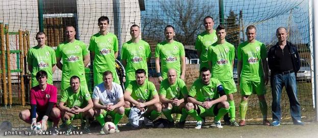 Gruppe Von Männern In Grünen T-Shirts Stehen Auf Einem Fussballfeld Mit Einem Ball Vor Ihnen, Einige Hocken, In Der Nähe Von Einem Torpfosten Mit Bäumen, Gebäuden Und Fahrzeugen Im Hintergrund.