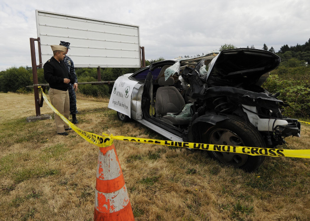 Polizist in Uniform neben einem schwer beschädigten Auto am Straßenrand mit eingedrückter Haube, zersplitterten Fenstern, einer Verkehrskegel, einem Band an einem Pfahl, Bäumen und einem bewölktem Himmel im Hintergrund.
