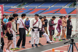 Eine Gruppe von Menschen steht auf einer Bahn mit einer Kamera auf einem Stativ rechts daneben, Stühle, Geländer und ein Banner im Hintergrund bei einem Sportereignis.
