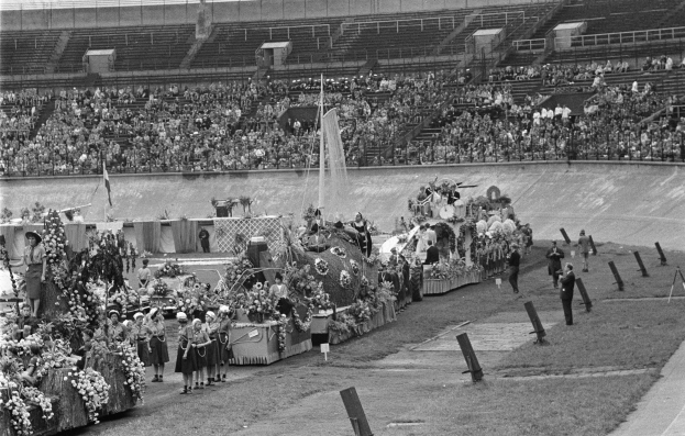 Schwarzes und weißes Foto eines Umzugs in einem Stadion mit Menschen, die stehen und sitzen, einem zentralen Springbrunnen und Blumenbouquets auf Fahrzeugen.