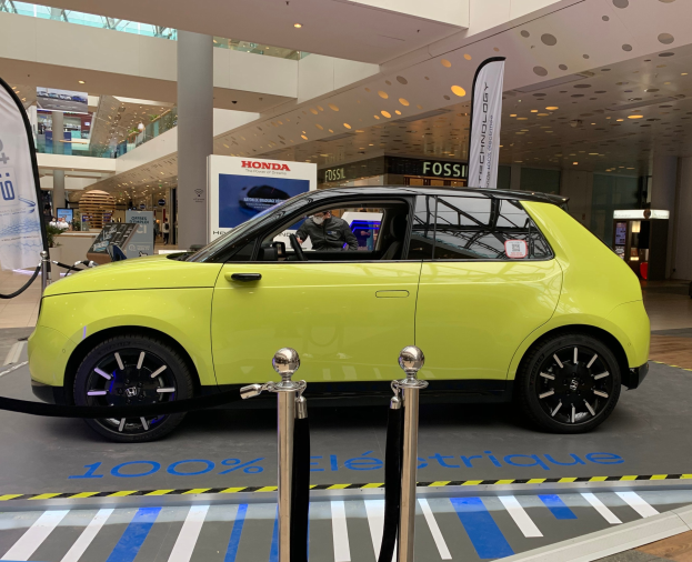 Ein gelbes wasserstoffbetriebenes Elektroauto vor einem Gebäude mit Absperrpollern, Bannern, Menschen, Schilden, einer Topfpflanze, Deckenleuchten und Ventilatoren.