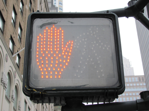 Rote Hand am Verkehrssignal in einer städtischen Umgebung mit Gebäuden auf beiden Seiten und einem klaren Himmel darüber.