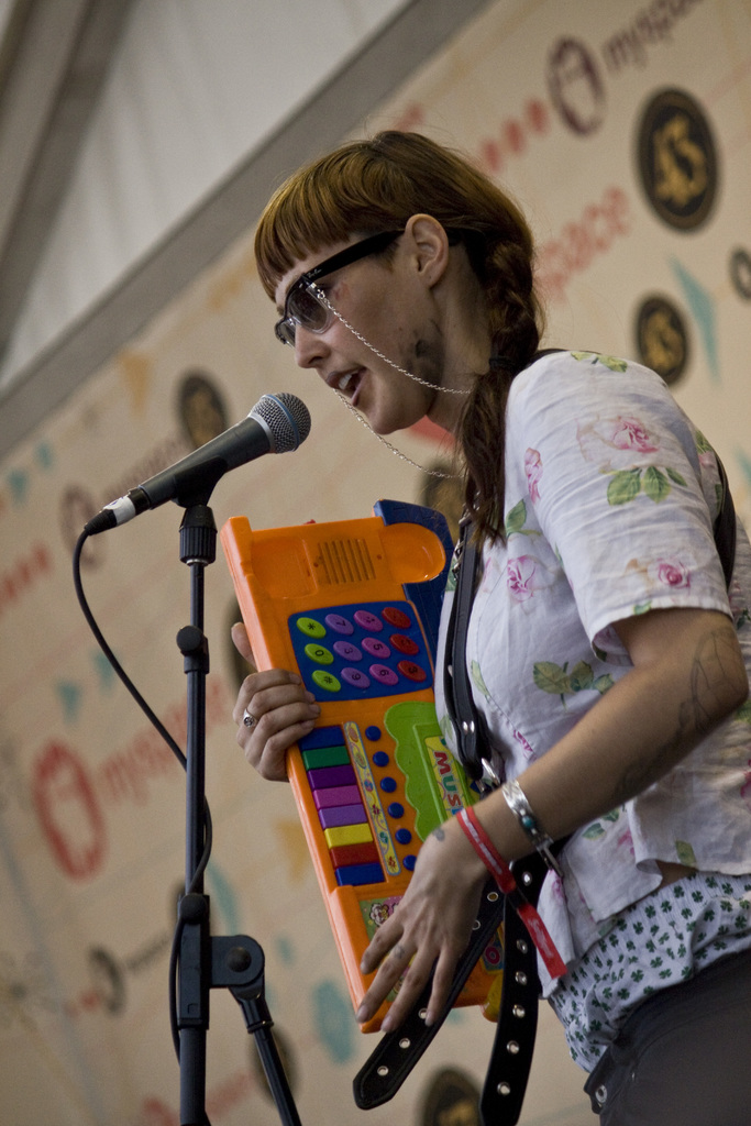 Eine Frau in einem wei├čen Hemd und Sonnenbrille singt in ein Mikrofon, w├Ąhrend sie eine Tastatur spielt, mit einem Plakat im Hintergrund.