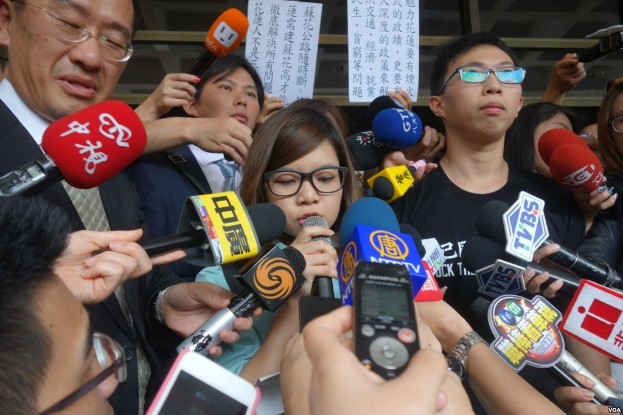 A group of people in a public space engaged in a heated discussion with reporters holding microphones, phones, and papers in the background.