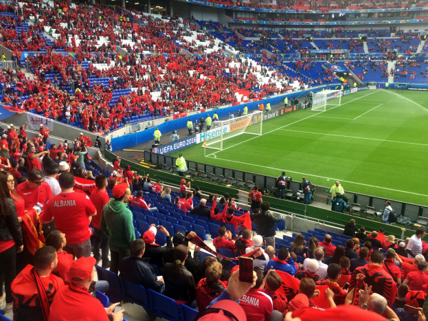 Eine große Menge an Menschen in einem Stadion schaut einem Fußballspiel zu, mit Menschen auf Sitzen und Stehplätzen, Bannern, einem Torpfosten und Deckenleuchten.