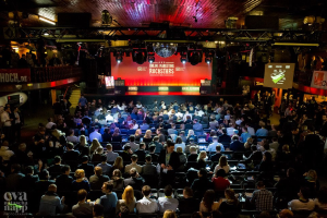 Eine große Menschenmenge sitzt auf Stühlen vor einer Bühne mit Bildschirmen und Bannern, beleuchtet von Scheinwerfern und umgeben von Geländern, voller Erwartung auf ein Online-Marketing-Rockstars-Event.