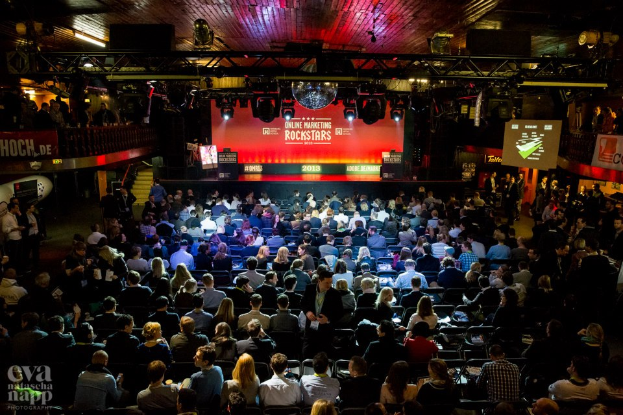 Eine große Menschenmenge sitzt auf Stühlen vor einer Bühne mit Bildschirmen und Bannern, beleuchtet von Scheinwerfern und umgeben von Geländern, voller Erwartung auf ein Online-Marketing-Rockstars-Event.