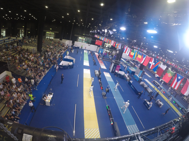 Große Halle mit Zuschauern bei einer Gymnastikveranstaltung, auf beiden Seiten sitzend und stehend auf dem Boden, unter Deckenlampen mit Flaggen im Hintergrund.