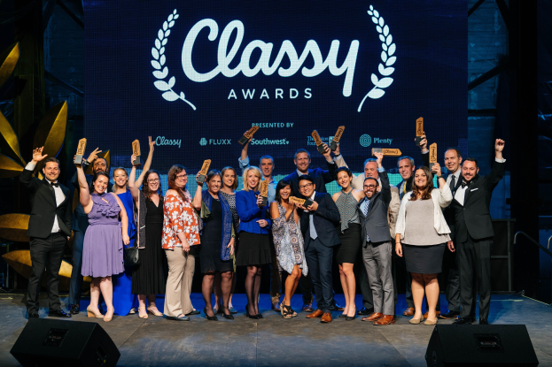 Gruppe von Menschen auf einer Bühne, die Auszeichnungen halten, mit Lautsprechern darunter und einem Hintergrundbild mit der Aufschrift "Classy Awards 2019."