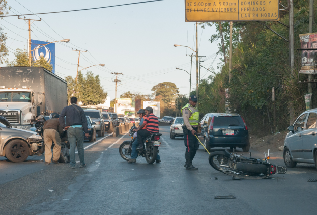 Gruppe von Menschen um ein verunglücktes Motorrad auf der Seite einer Straße mit mehreren Fahrzeugen, Bäumen, Pfählen, Lampen, Schildern und dem Himmel im Hintergrund.