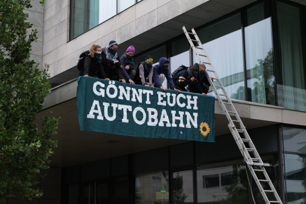 Eine Gruppe von Menschen auf einem Gebäude, das eine Fahne hält, mit einem Baum auf der linken Seite und Glasfenstern im Hintergrund, die scheinbar für die deutsche Regierung protestieren.