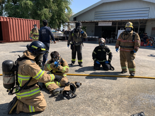 Feuerwehrleute mit Helmen und Gasmasken sitzen auf dem Boden während einer Übung vor einem Gebäude, mit einem Rohr im Vordergrund, einem Container links, Bäumen und einem Schuppen im Hintergrund und Himmel darüber.