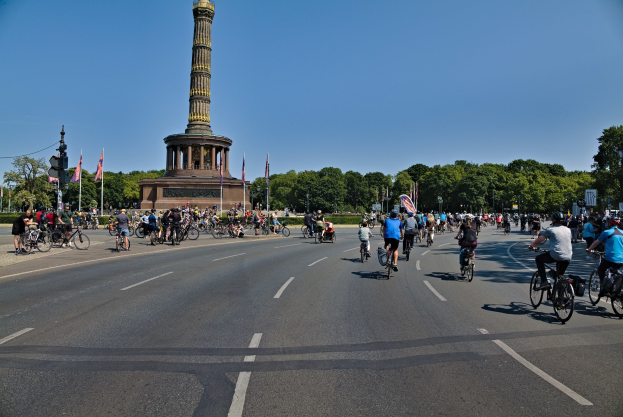 Eine große Gruppe von Menschen auf Fahrrädern fährt eine Straße entlang, mit einem Denkmal im Hintergrund, das von Fahnenmasten und Bäumen umgeben ist und einen sichtbaren Himmel hat.